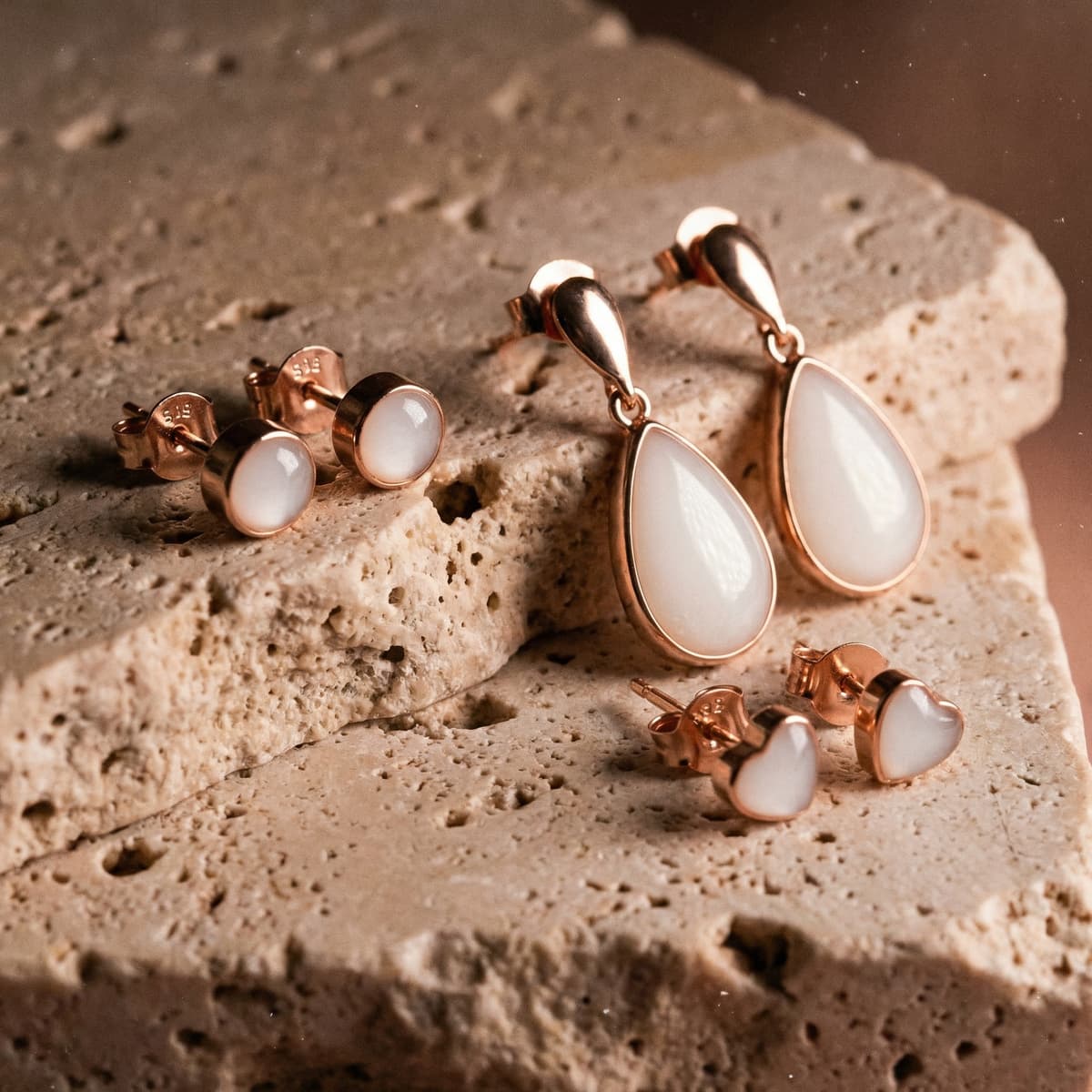 Juego de joyería en oro rosa con aretes leche materna, piedras blancas en forma de gota, corazón y stud redondo sobre fondo de piedra natural.