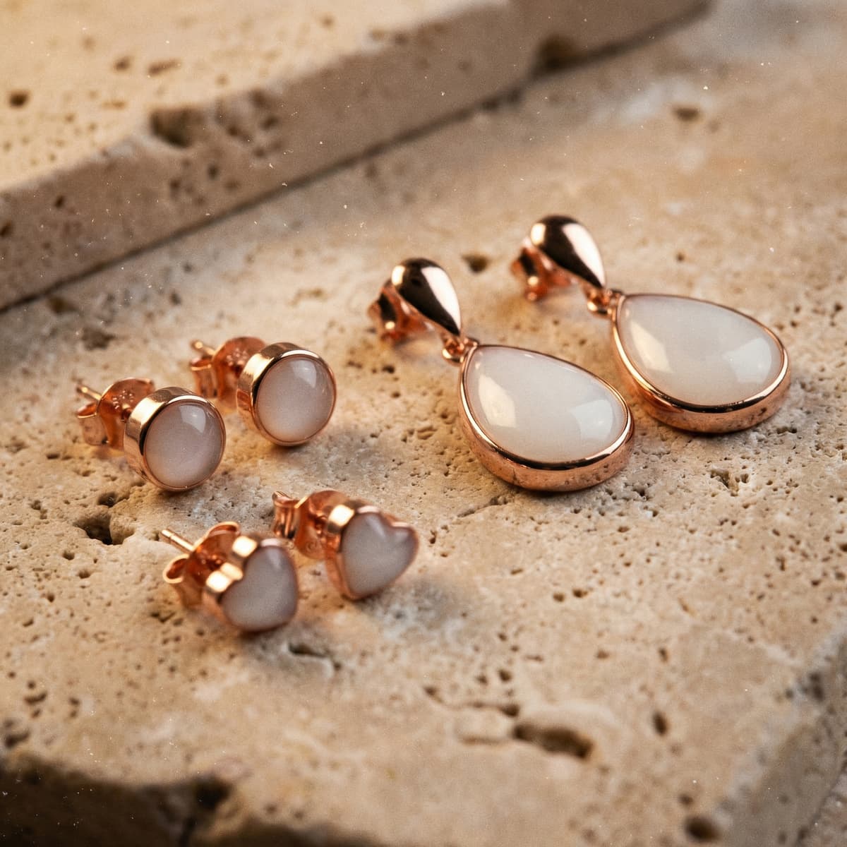 Set de aretes en tono oro rosa con piedras blancas y diseños de gota, redondo y corazón sobre fondo de piedra natural, ideal para aretes leche materna diy.