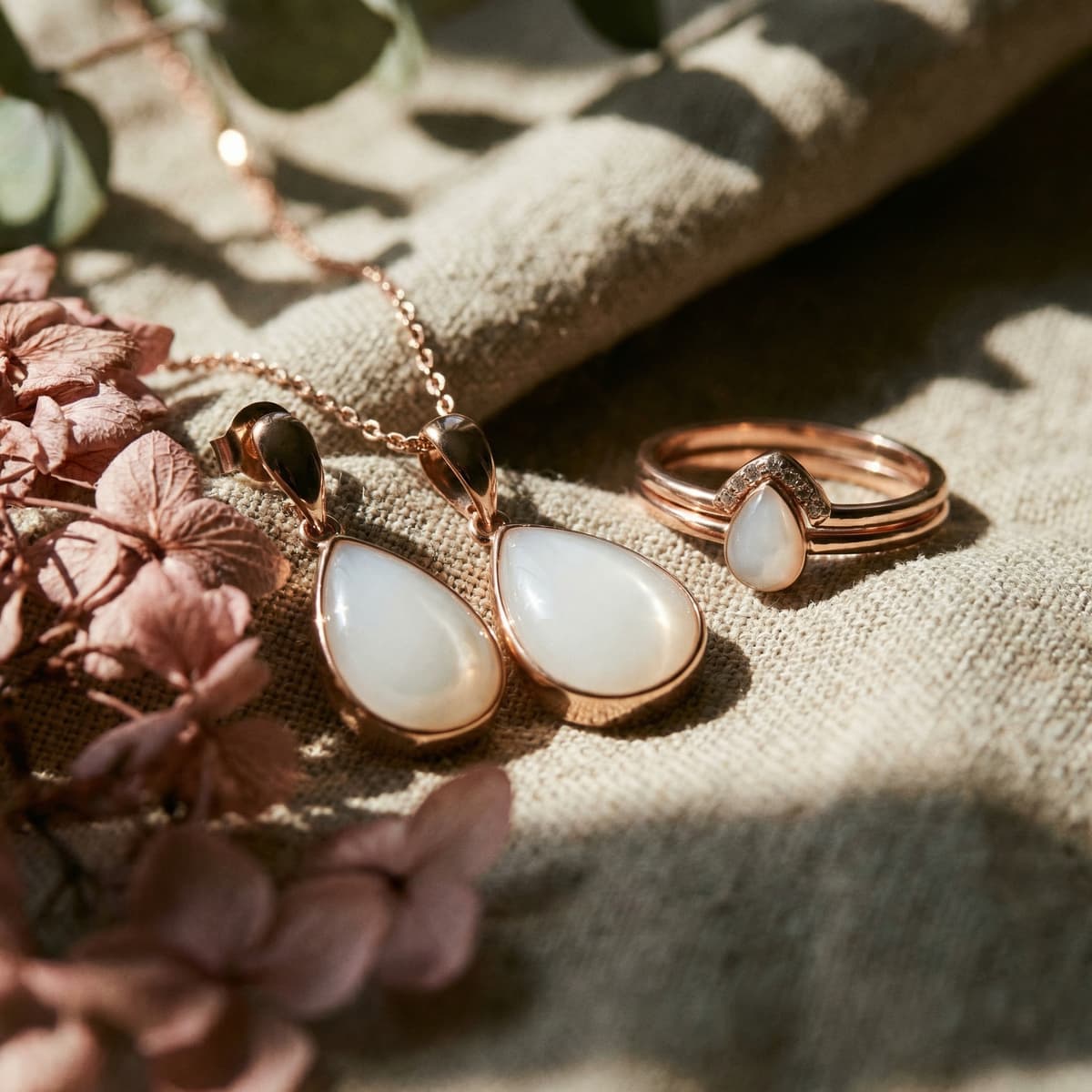 Joyas conmemorativas para el destete: collar con colgante en forma de lágrima y anillo tono oro rosa con piedra blanca sobre tela beige con flores secas