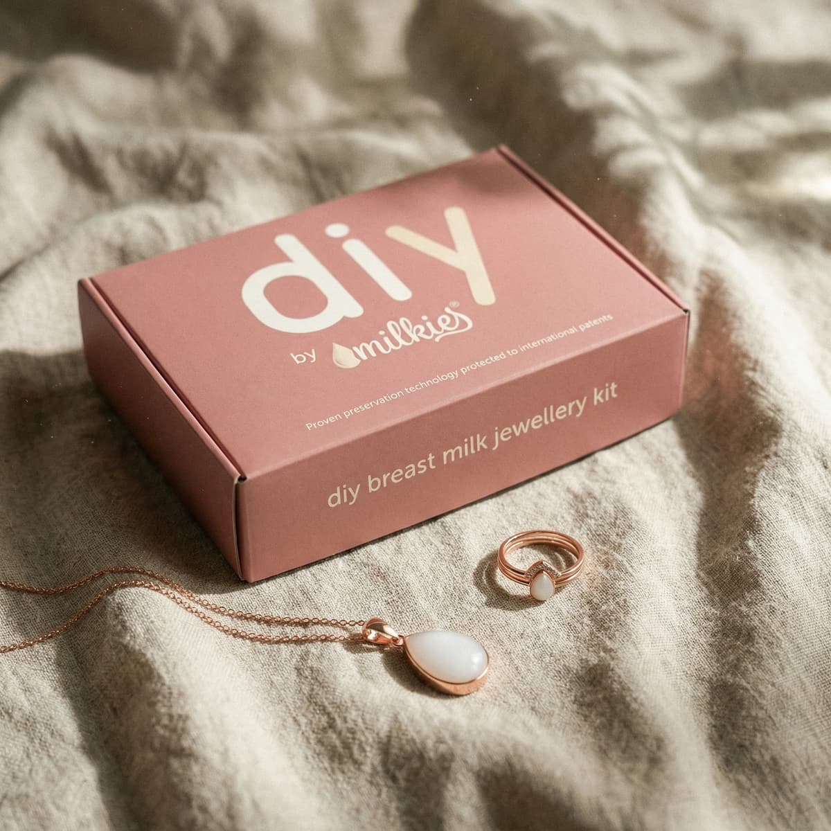 Caja rosa del kit DIY by MILKIES de joyería con leche materna, con collar y anillo en tono oro rosa sobre tela beige, como ideas regalo nacimiento para mamás.