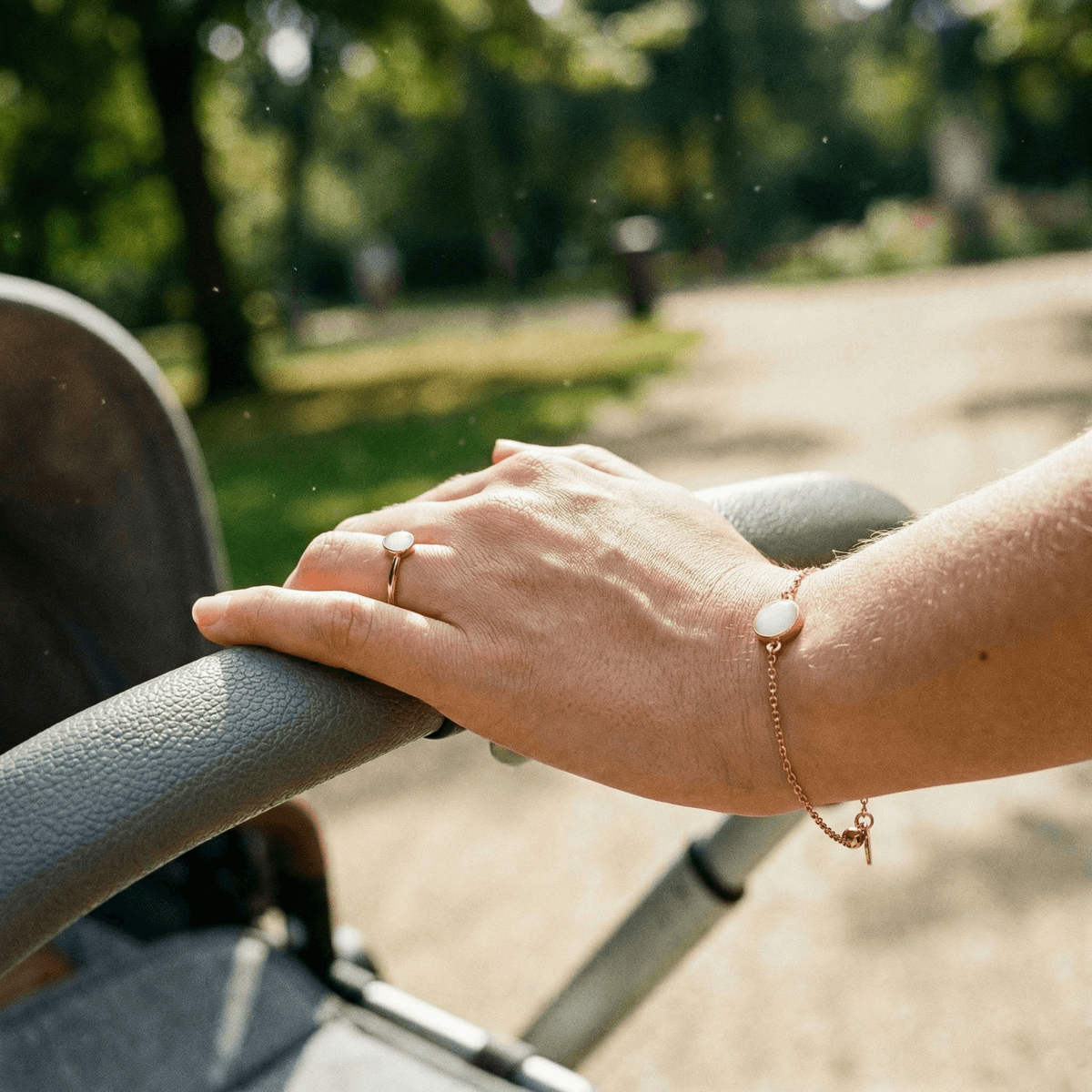 Mano de una madre empujando un cochecito en un parque, luciendo joyas de leche materna: anillo y pulsera dorados con piedra blanca.