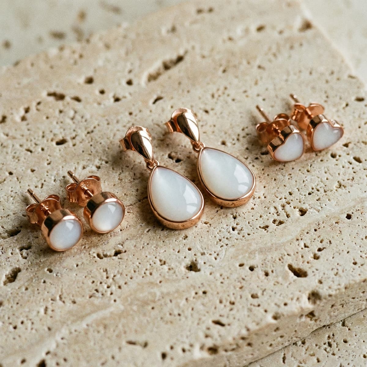 Conjunto de joyas pendientes leche materna en oro rosa, con diseño de gota y botón, resina blanca tipo piedra sobre fondo de piedra beige texturizada.