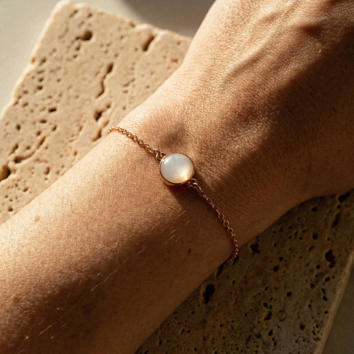 Pulsera minimalista de cadena fina en tono oro rosa con cabujón blanco tipo opalina, ideal para joyas pulsera leche materna, sobre muñeca iluminada con luz cálida y fondo de piedra natural.