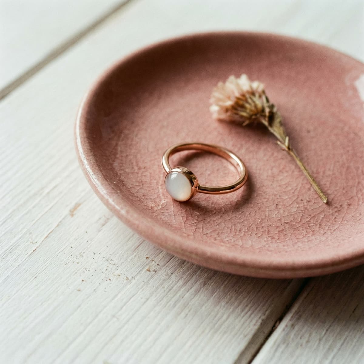 Anillo dorado con piedra blanca tipo cabujón sobre un plato rosa junto a una flor seca, ideal para un kit anillo leche materna de recuerdo.