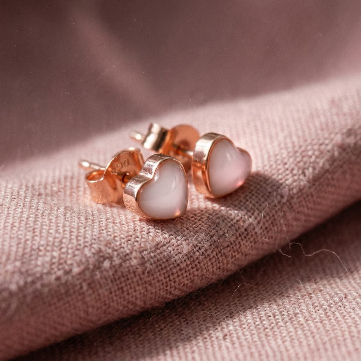 Aretes tipo botón en forma de corazón tono oro rosado con centro blanco lechoso, ejemplo de kit aretes leche materna para crear un recuerdo en casa sobre fondo de tela rosa.