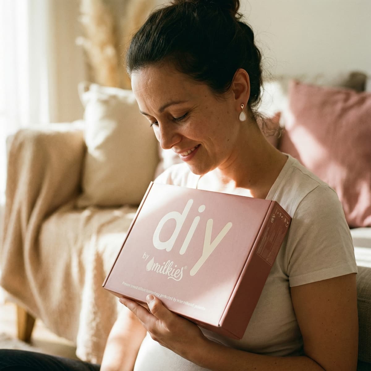 Madre sonriente en casa sosteniendo una caja “DIY by MILKIES”, mostrando por qué el kit creación pendientes leche es una opción práctica y personal para transformar la leche materna en una joya recuerdo.