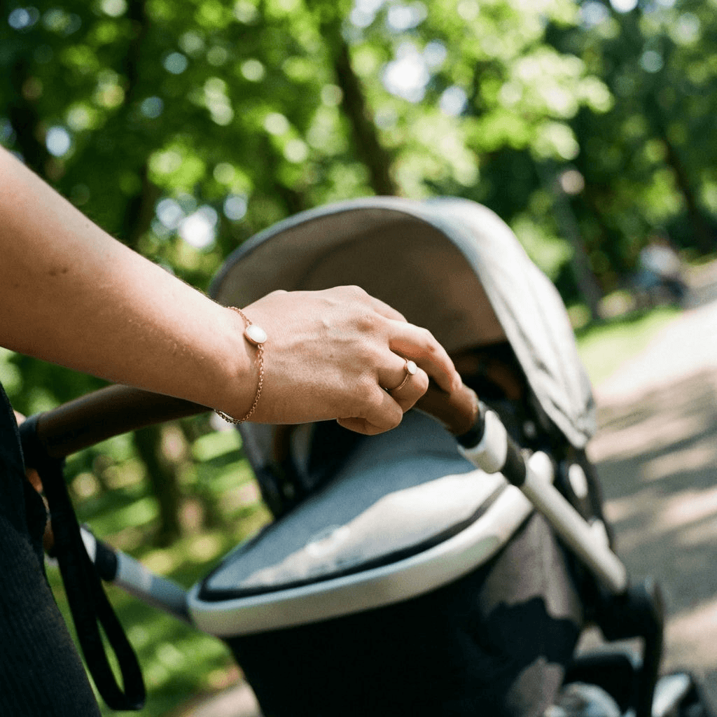Madre empujando un cochecito de bebé en un parque, luciendo un anillo y una pulsera dorada tipo kit pulsera lactancia con piedra blanca.
