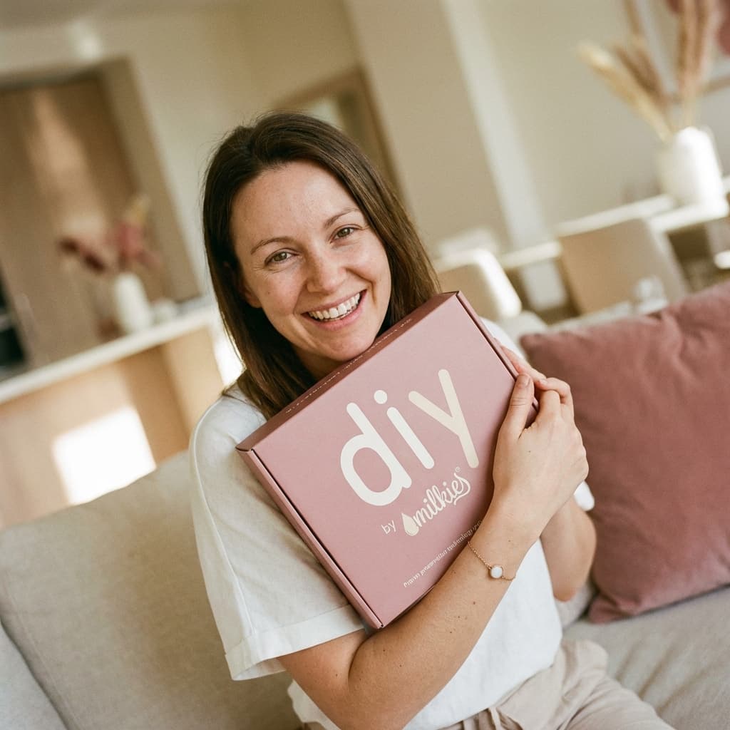 Madre sonriendo en casa sosteniendo la caja rosa DIY by MILKIES, mostrando por qué elegir un kit pulsera leche materna es una opción práctica para crear un recuerdo personal en privado.