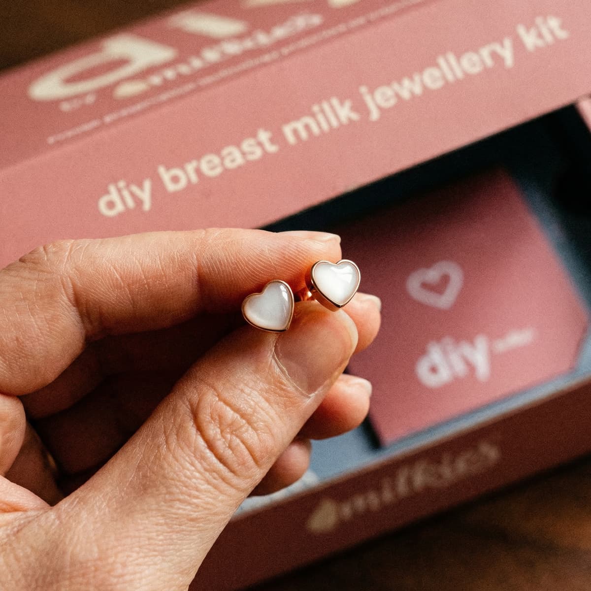 Mano sosteniendo delicados pendientes leche materna en forma de corazón, con acabado dorado y centro blanco, frente a la caja del kit DIY breast milk jewellery de MILKIES.