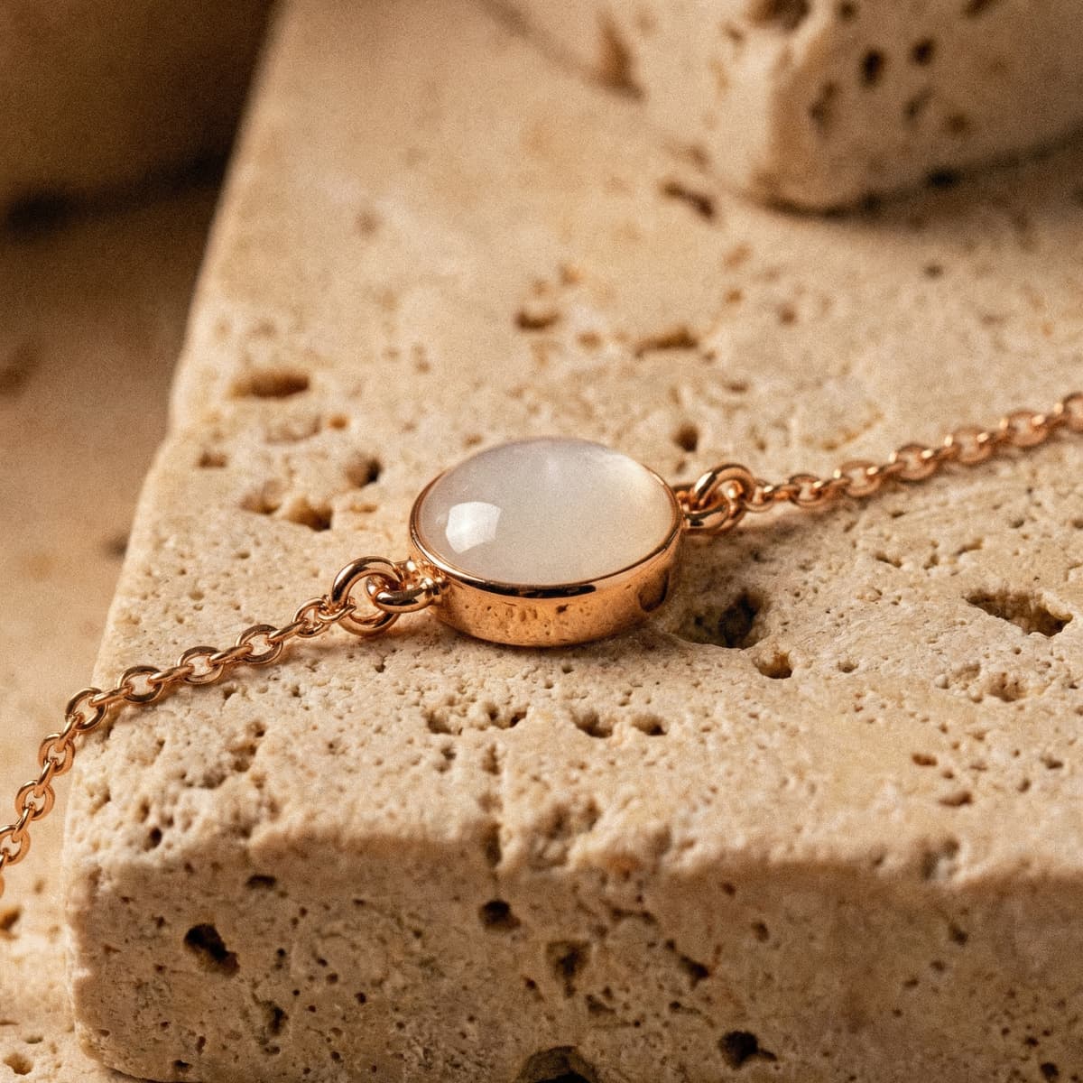 Primer plano de una pulsera leche materna con cadena y engaste en oro rosa, con piedra ovalada blanca translúcida sobre una superficie de piedra beige.