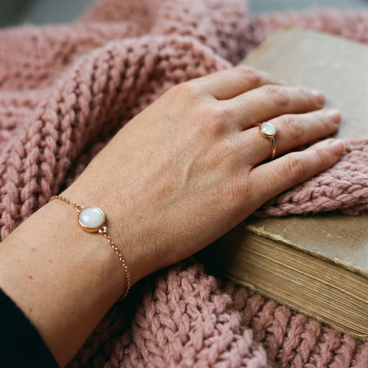 Mano sobre manta de punto rosa mostrando anillo y pulsera de oro rosa con piedra blanca, un delicado recuerdo fin lactancia en joyería minimalista junto a un libro antiguo.