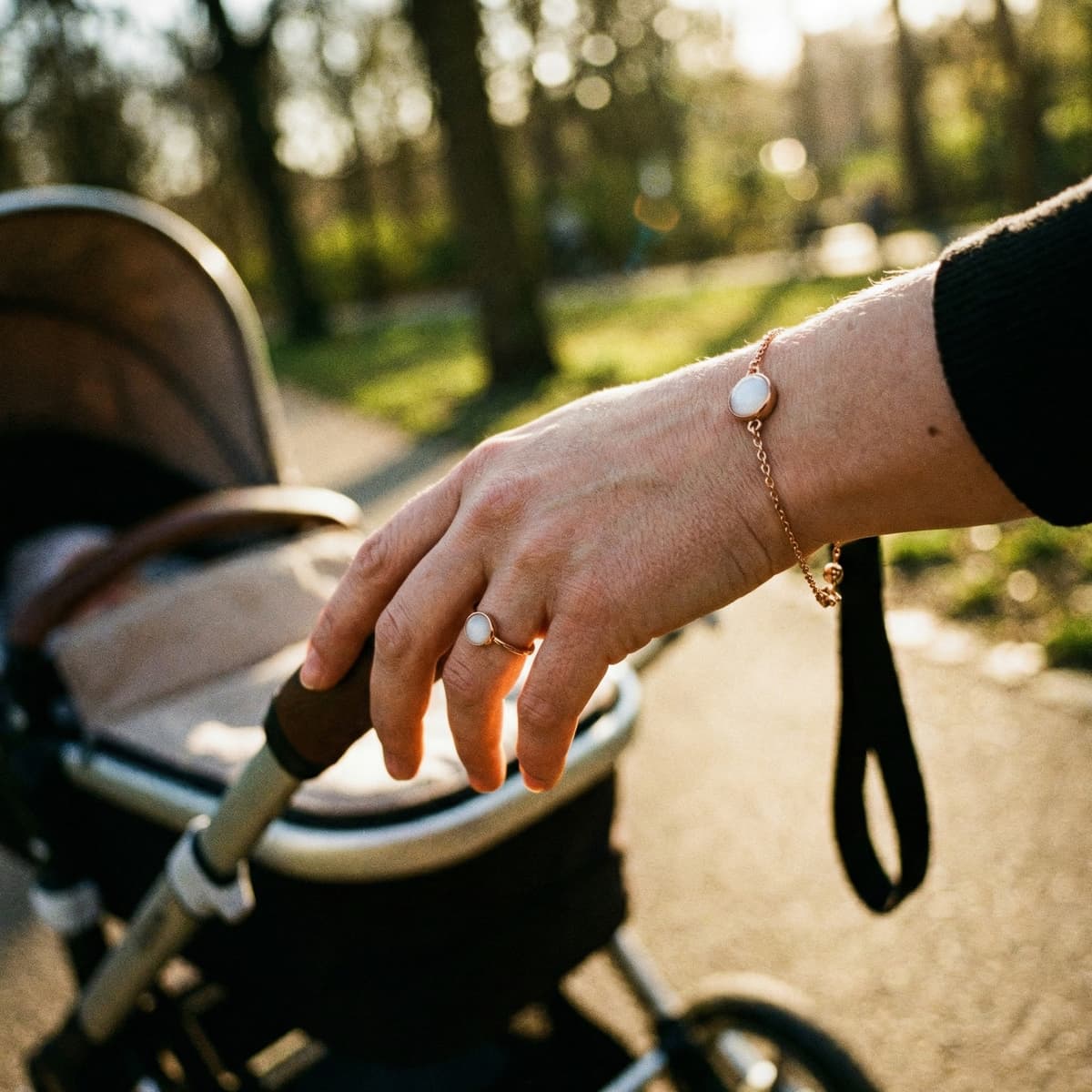 Madre empujando un cochecito en un paseo al aire libre con luz cálida, luciendo pulsera y anillo con piedra blanca tipo recuerdo leche materna en la mano.