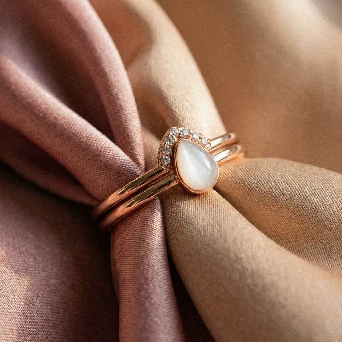 Anillo de oro rosa con piedra en forma de lágrima y pequeños cristales sobre tela satinada rosa y beige, joya elegante y delicada como recuerdo maternidad.