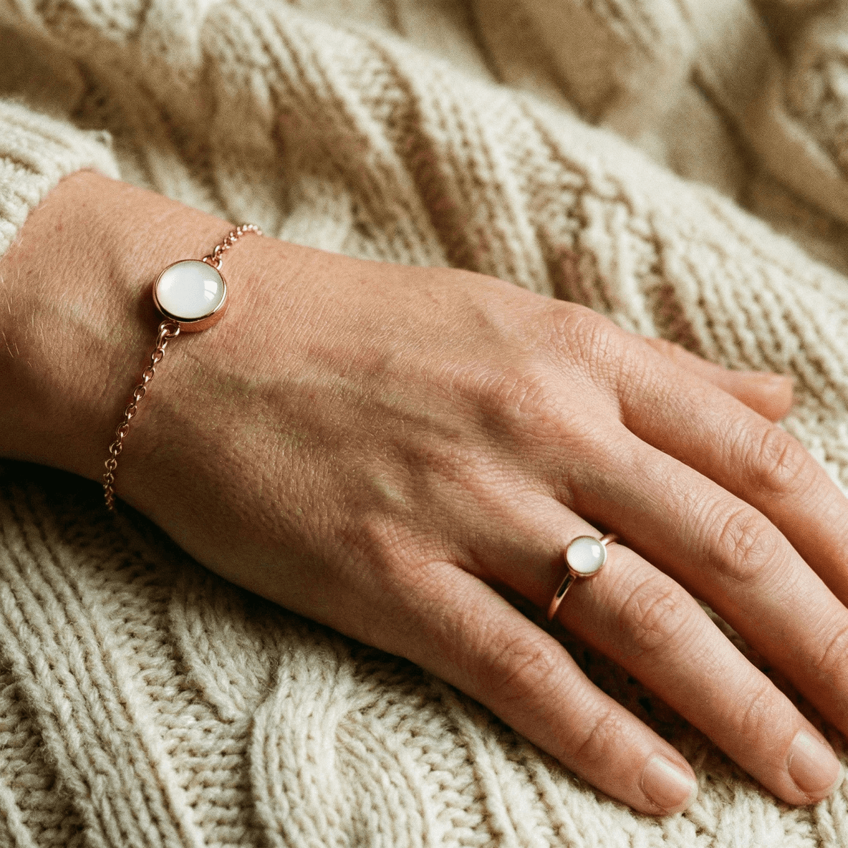 Mano sobre manta de punto beige mostrando anillo y pulsera dorados con piedra blanca, joyas delicadas ideales como regalo destete para mamás.