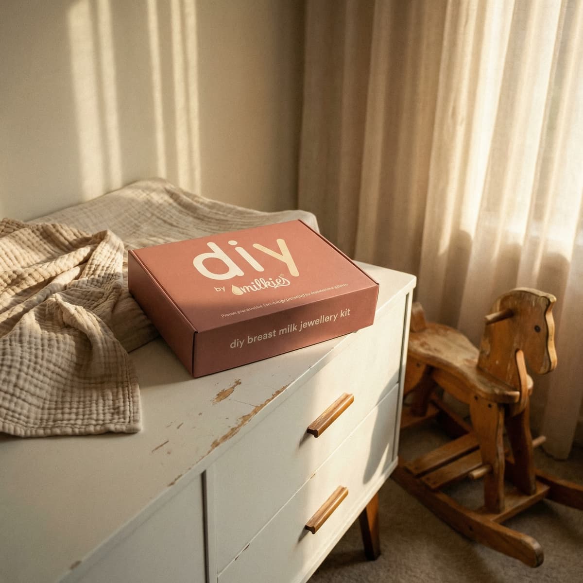 Caja del kit DIY by MILKIES para crear joyería de leche materna, un regalo embarazada original, sobre una cómoda blanca en habitación infantil con luz cálida y caballito de madera.