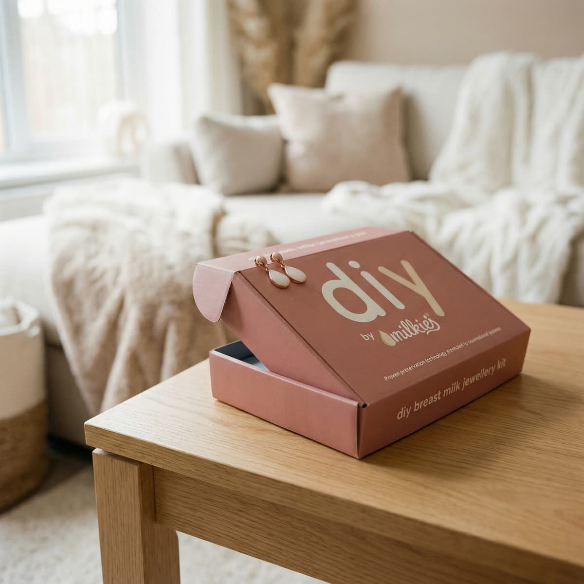 Kit DIY by MILKIES para crear joyería con leche materna, presentado como regalo fin de lactancia en una caja rosa sobre una mesa de madera en un salón acogedor con pendientes.