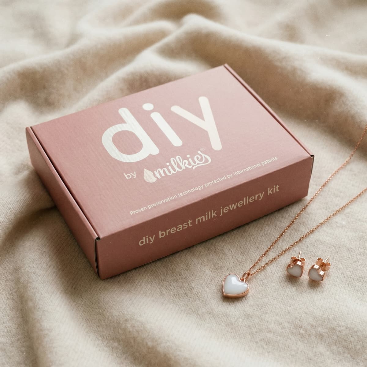 Kit DIY by MILKIES para joyería de leche materna, un regalo madre primeriza único: caja rosa con collar y pendientes de corazón en tono oro rosa sobre tela beige.