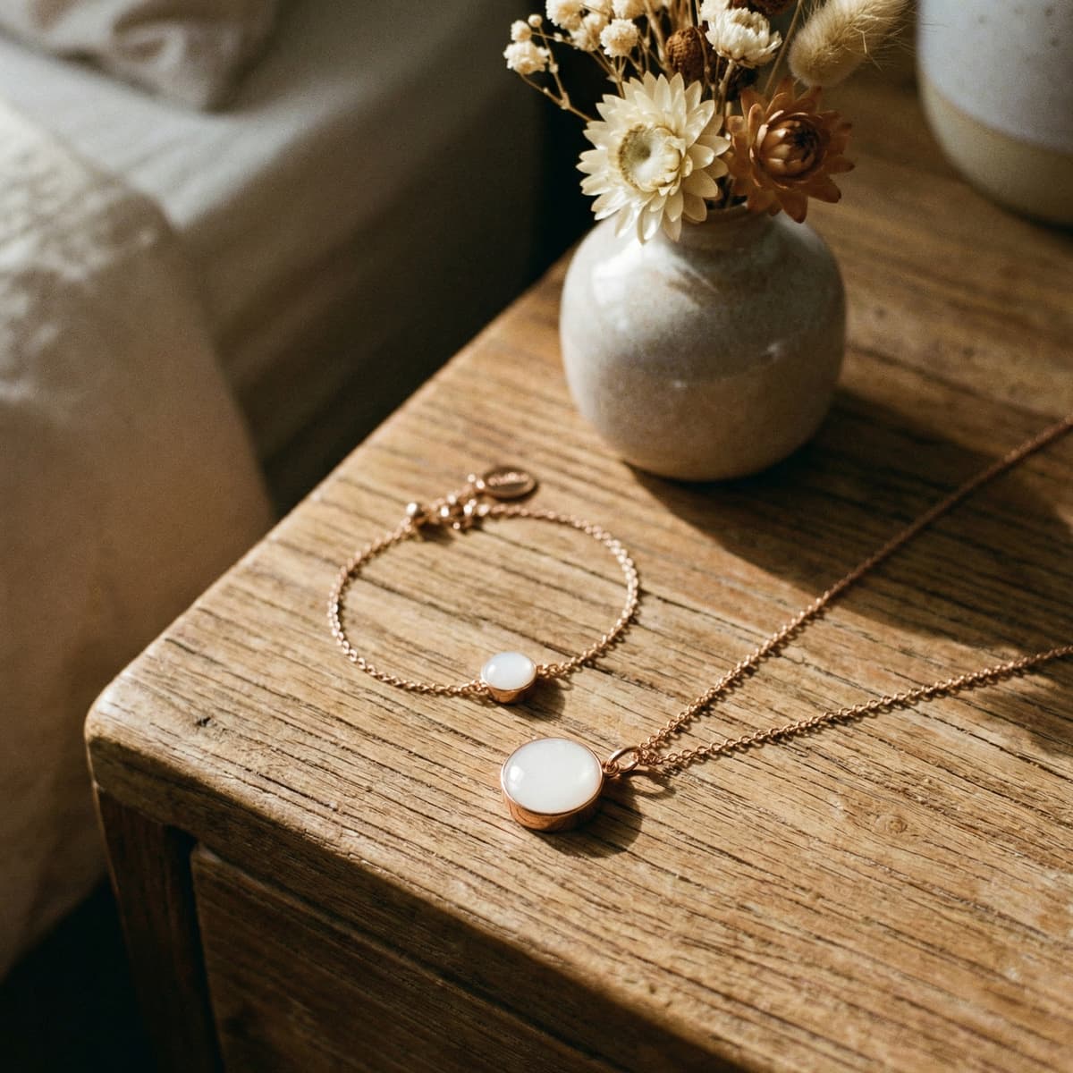 Joyas minimalistas en tono oro rosa con piedra blanca tipo leche materna sobre mesa de madera junto a un jarrón con flores secas, ideal como regalo nacimiento bebe elegante y sentimental.