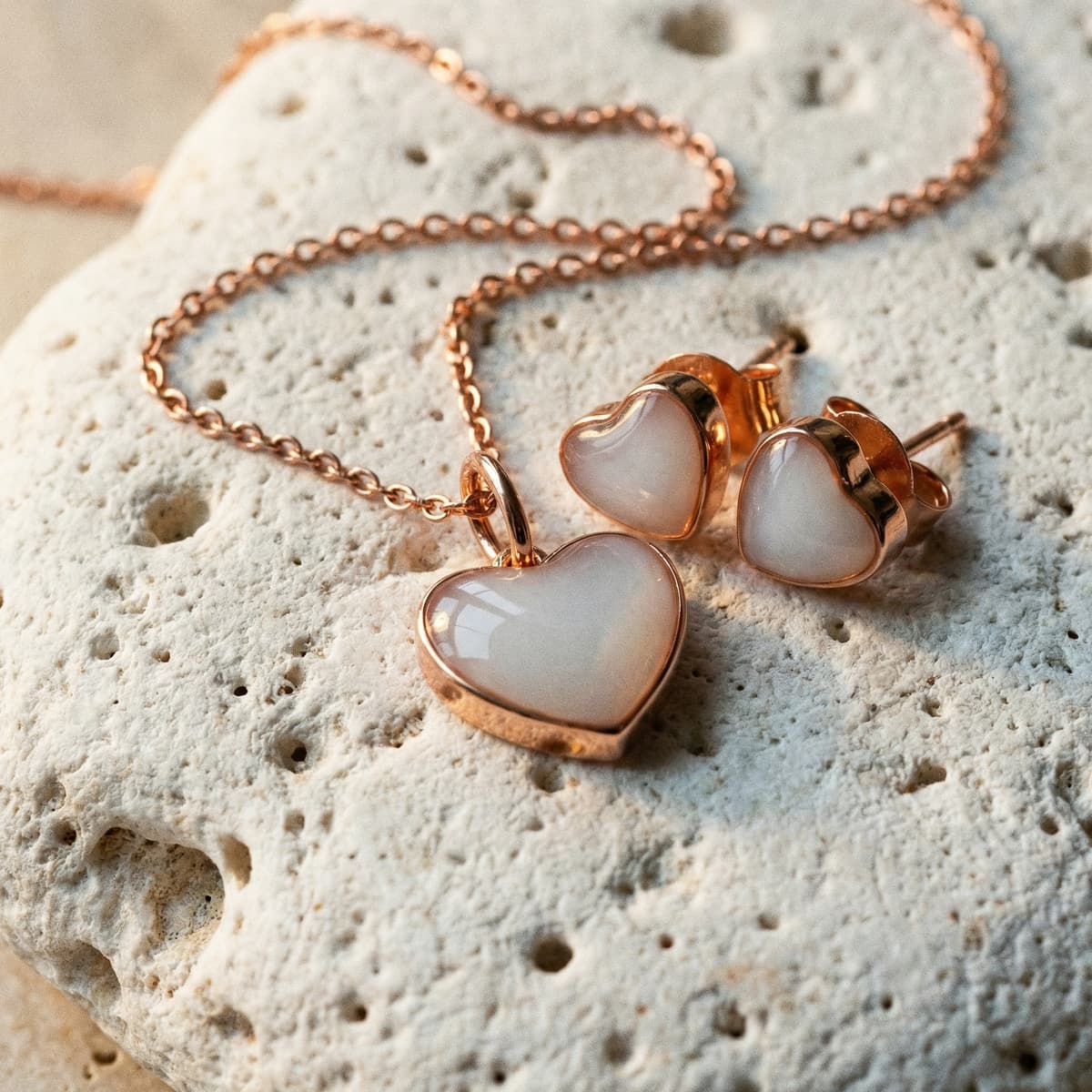 Conjunto de joyas en oro rosa con colgante y pendientes en forma de corazón con piedra blanca sobre fondo de piedra, ideal como regalo original nacimiento para mamá.