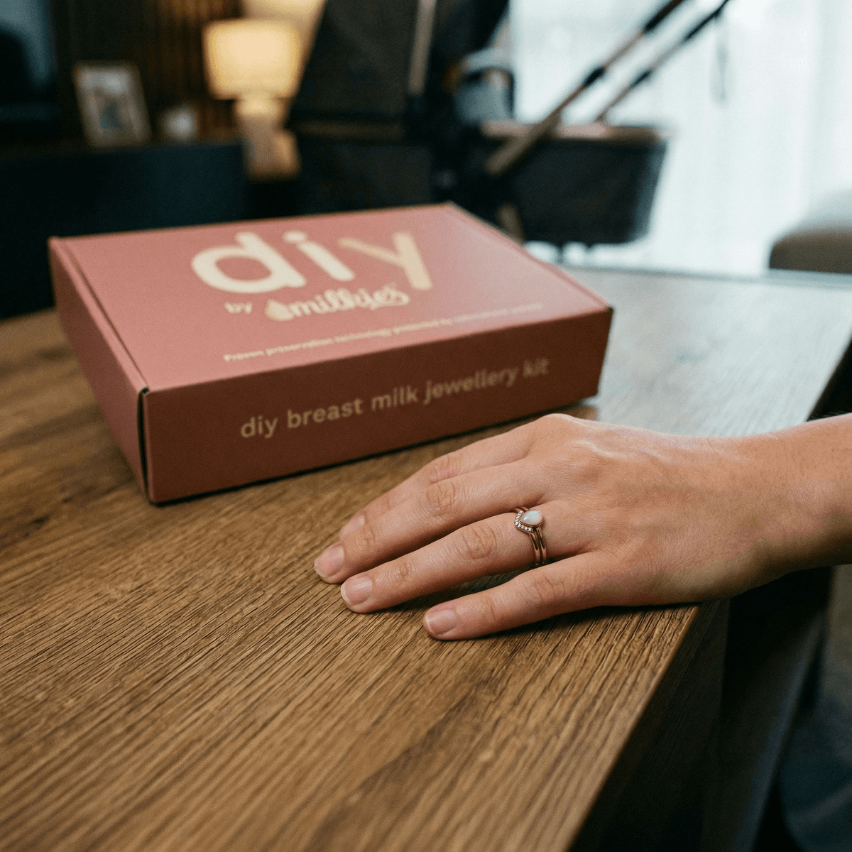 Kit DIY by MILKIES para crear joyería con leche materna en casa, ideal como regalo original nacimiento; caja del kit sobre una mesa y mano mostrando un anillo con piedra blanca.