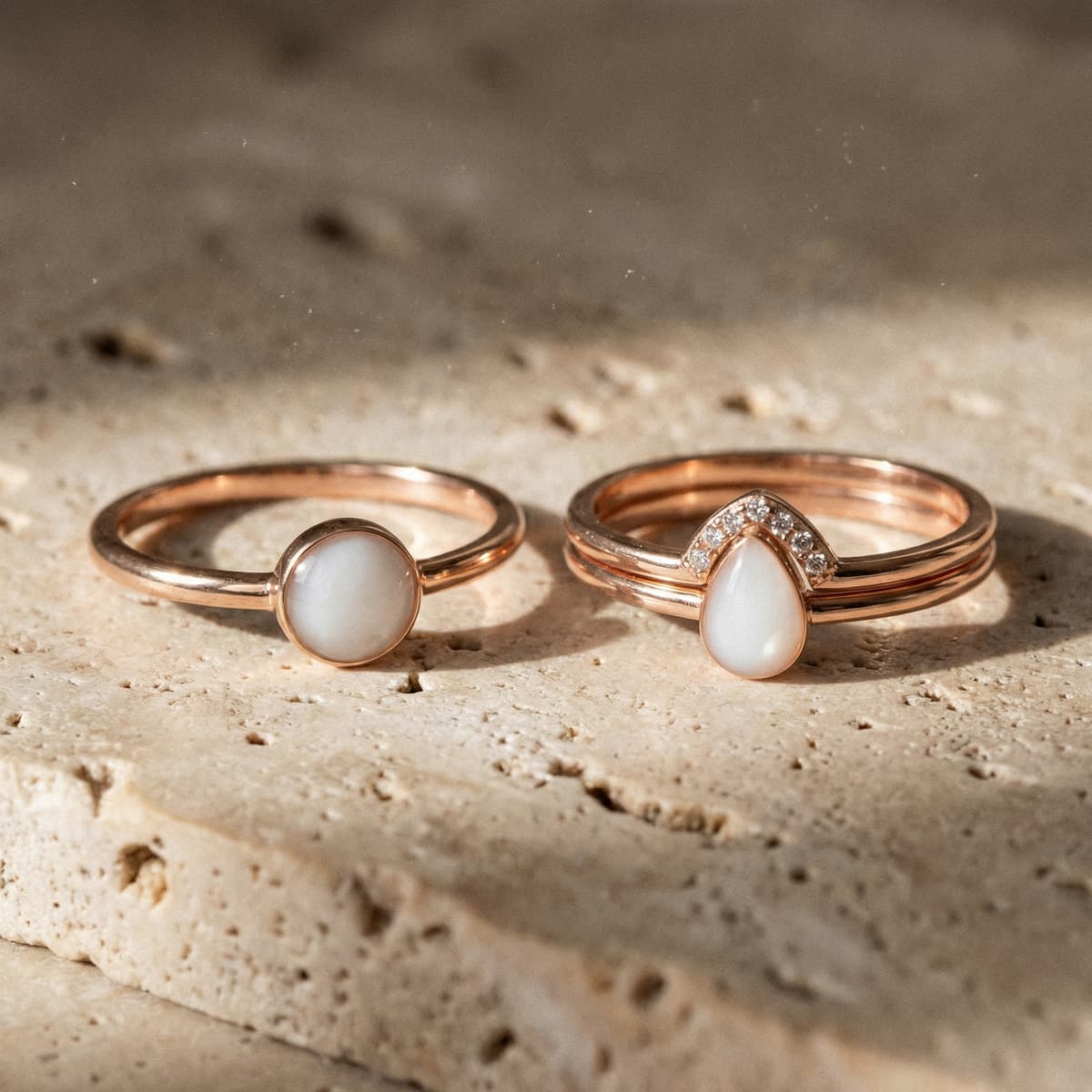 Dos anillos finos color oro rosa con piedra blanca tipo leche materna (uno redondo y otro en forma de gota con detalles brillantes) sobre fondo de piedra natural, ideal como set anillo leche materna.