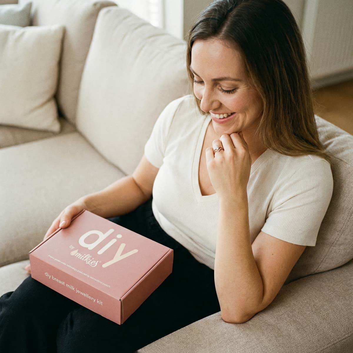 Mamá sonriente en el sofá con una caja rosa del kit DIY by MILKIES, una buena idea para crear en casa un set anillo leche materna y otros recuerdos personalizados con privacidad.