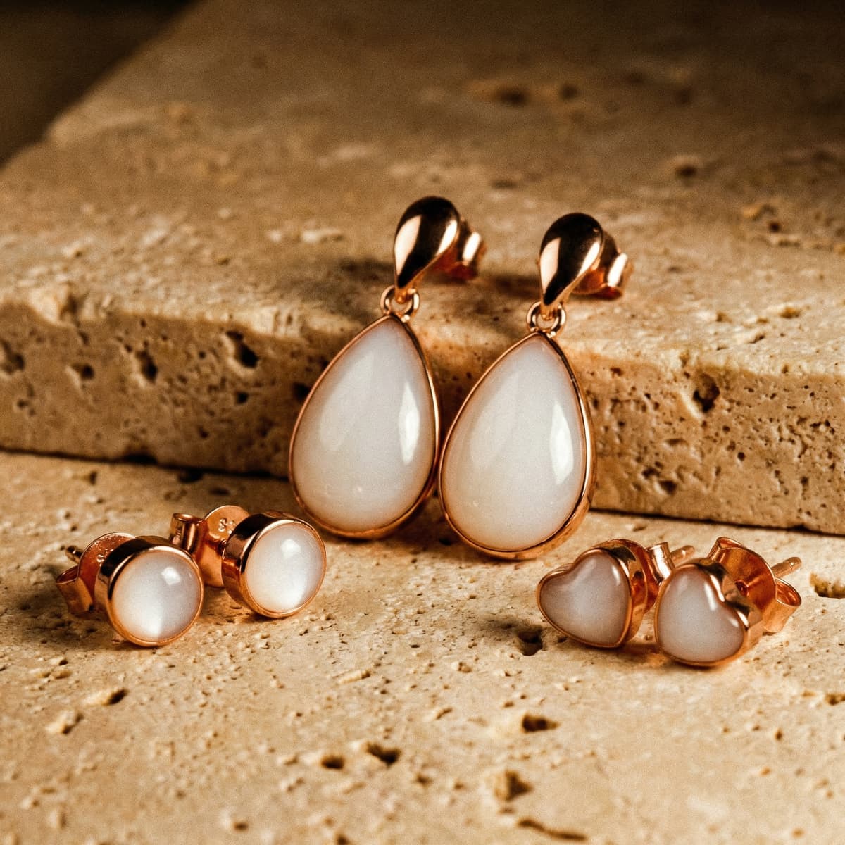 Elegante set pendientes leche materna en tono oro rosa con gotas colgantes y studs redondos y en forma de corazón sobre fondo de piedra beige.