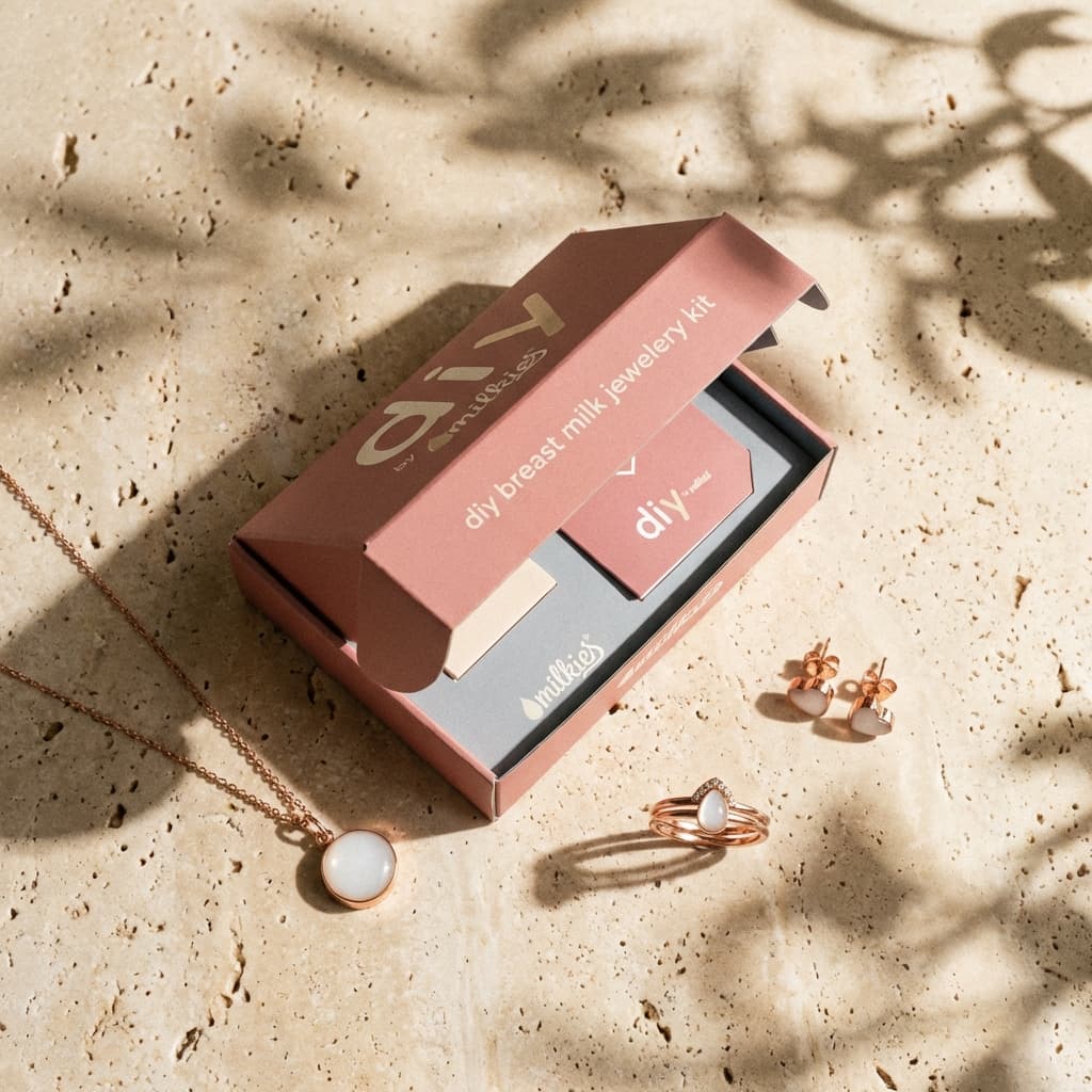 Caja rosa abierta de kit creación joyas leche materna DIY by MILKIES sobre fondo beige, con colgante, anillo y pendientes en tono oro rosa dentro del kit.