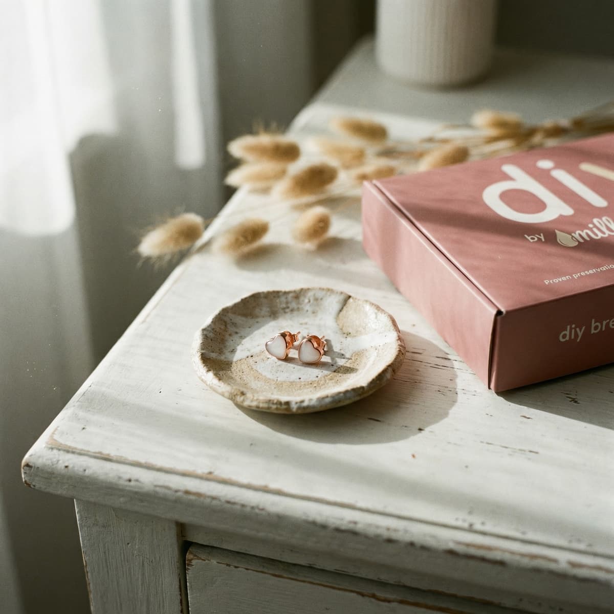 Estuche DIY by MILKIES en caja rosa junto a un plato con pendientes tipo botón blancos y montura dorada, ideal como kit hacer pendientes leche materna, sobre mesa de madera con luz natural.