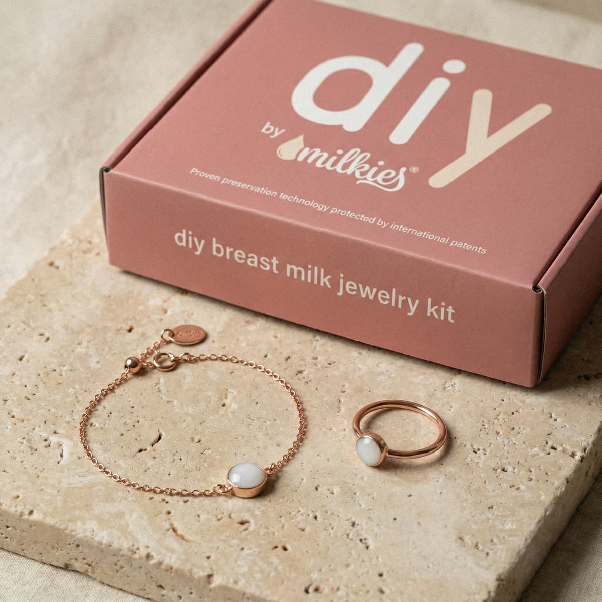 Kit DIY by MILKIES con caja rosa para kit hacer pulsera leche materna en casa, junto a pulsera y anillo dorados con piedra blanca de resina para conservar leche materna.