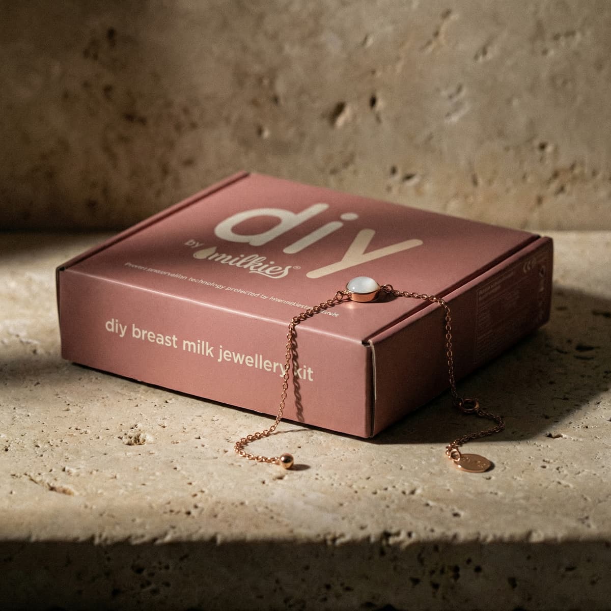 Caja rosa de DIY by Milkies con el texto “diy breast milk jewellery kit” y una pulsera de cadena dorada con piedra blanca encima, ideal como kit hacer pulsera leche materna en casa.