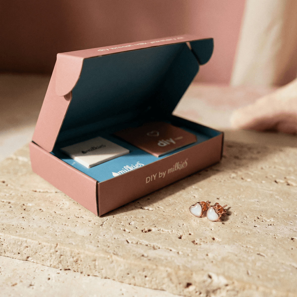 Caja abierta de DIY by MILKIES con el contenido del kit joyería pendientes lactancia y unos pendientes de corazón con acabado dorado sobre una superficie de piedra.