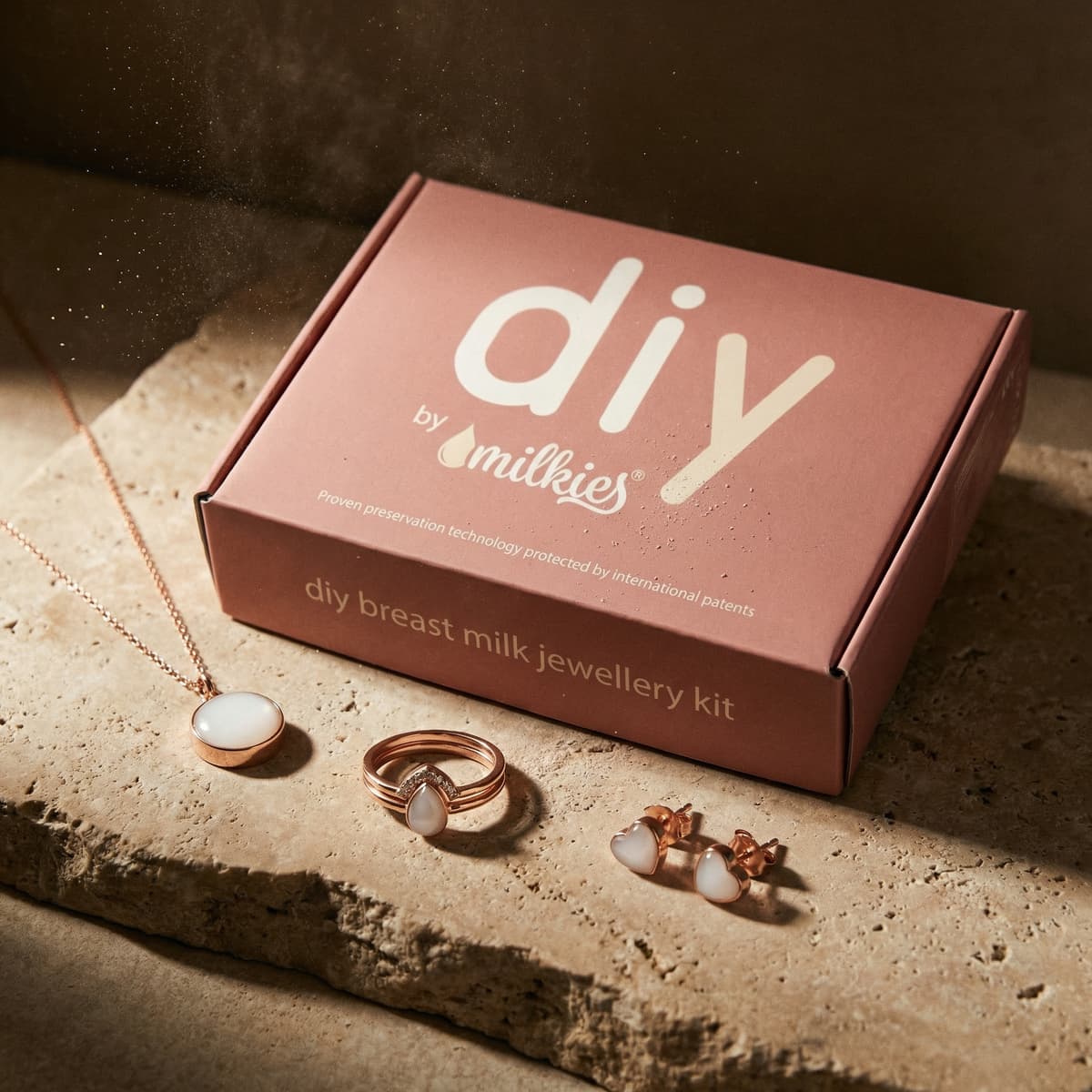 Caja rosa de DIY by MILKIES con un kit para hacer joyas de leche materna, junto a un collar, un anillo y pendientes en tono oro rosa con piedra blanca sobre una superficie de piedra.