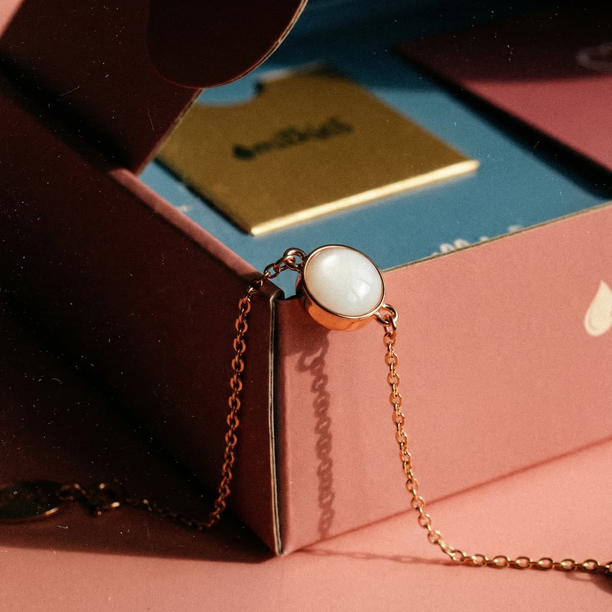 Kit de joyería DIY con materiales hacer pulsera leche: cadena dorada con cabujón blanco tipo leche materna sobre caja rosa, en fondo de papelería azul.