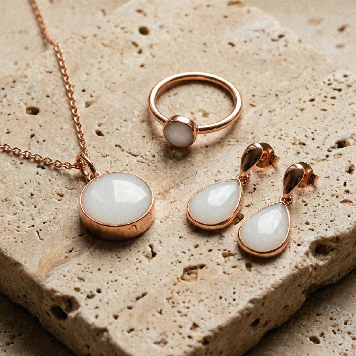 Conjunto de joyas en tono oro rosa con colgante redondo, anillo y pendientes tipo lágrima con piedra blanca de resina para leche materna sobre fondo de piedra